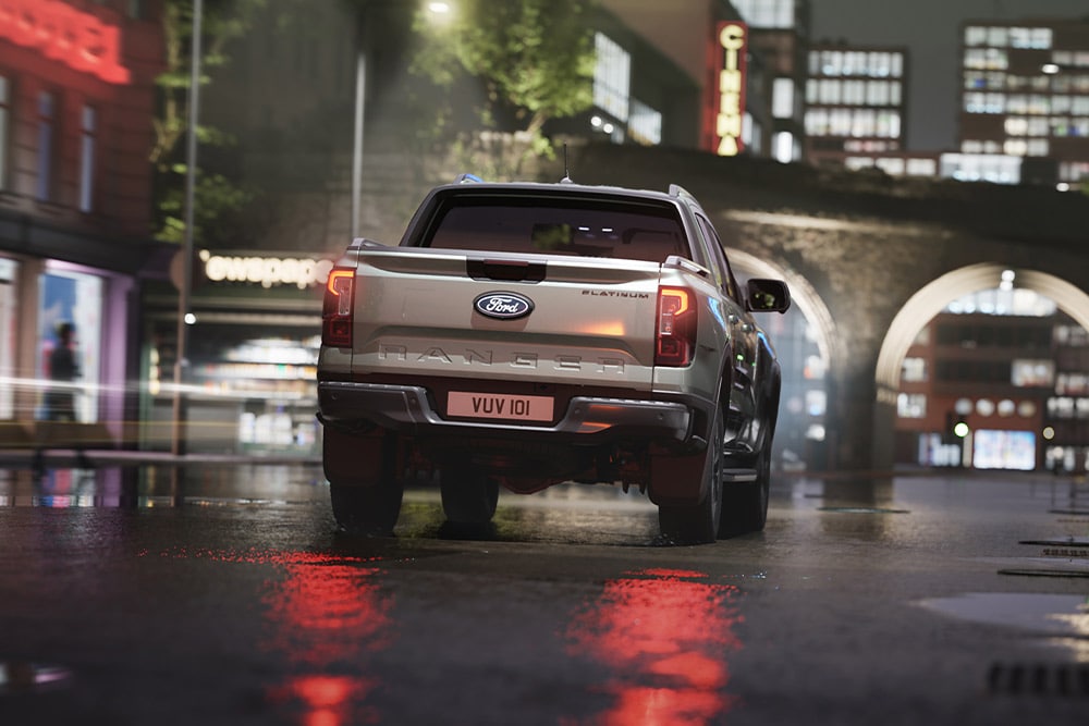 Vista trasera de una Ford Ranger plateada circulando por un paisaje urbano iluminado con luces de neón por la noche.