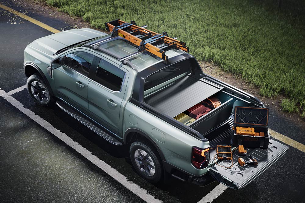 Vista trasera en ángulo alto de una camioneta Ford Ranger gris, con la caja abierta con herramientas y una baca en una superficie pavimentada.