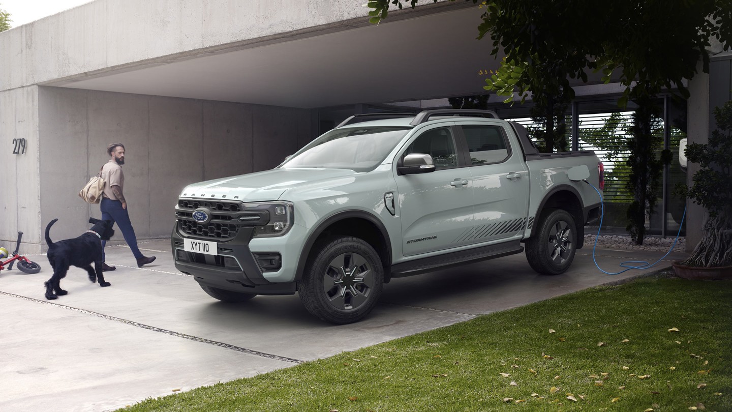 Un hombre y su perro entran en una casa moderna, pasando al lado de un Ford Ranger Stormtrak híbrido enchufable que está aparcado, cargando.