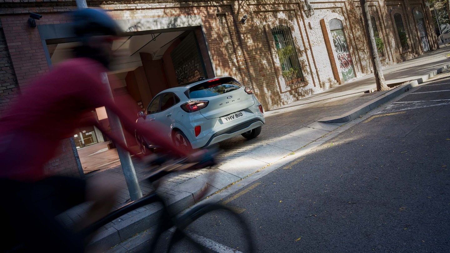 Ford Puma gris sale de una entrada, ciclista vestido de rojo pasa.