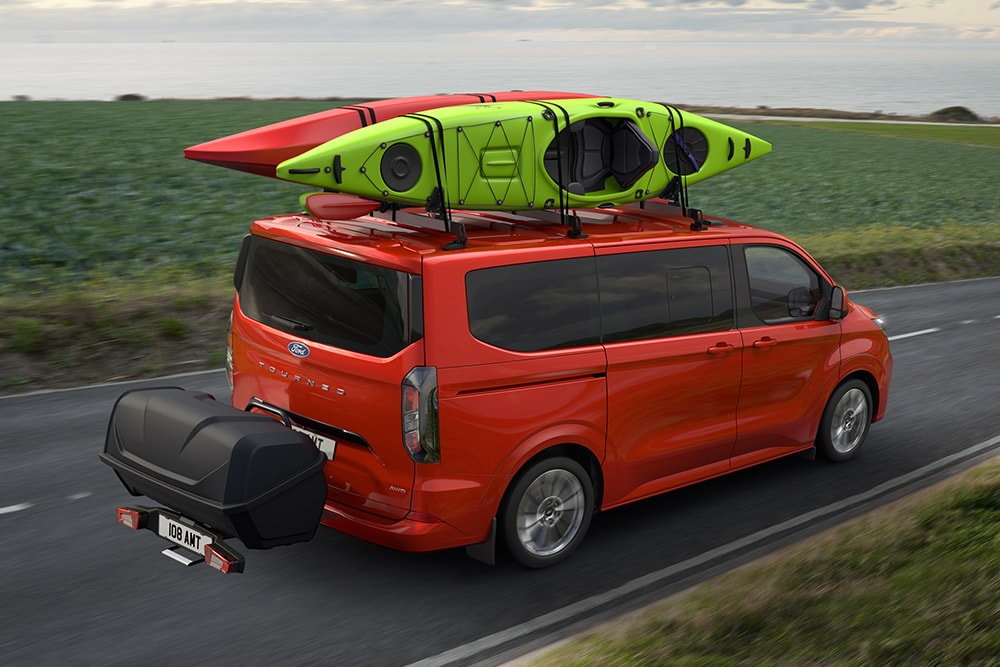 Ford Pro Transit rojo con dos kayaks en el techo, remolcando un portaequipajes y conduciendo por una carretera costera.