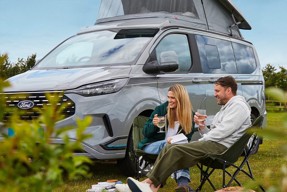 Una pareja disfrutando de bebidas en sillas de camping junto a una furgoneta camper Ford Transit Custom gris con techo elevable.