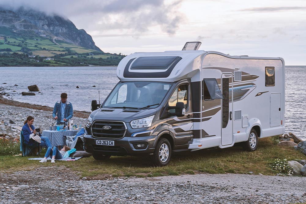 Una pareja disfrutando de bebidas en sillas de camping junto a una furgoneta camper Ford Transit Custom gris con techo elevable.