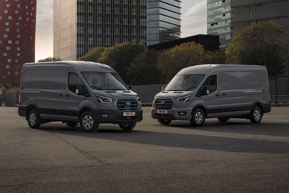 Dos furgonetas Ford Pro Transit, una de ellas eléctrica, aparcadas en un solar con edificios modernos al fondo.