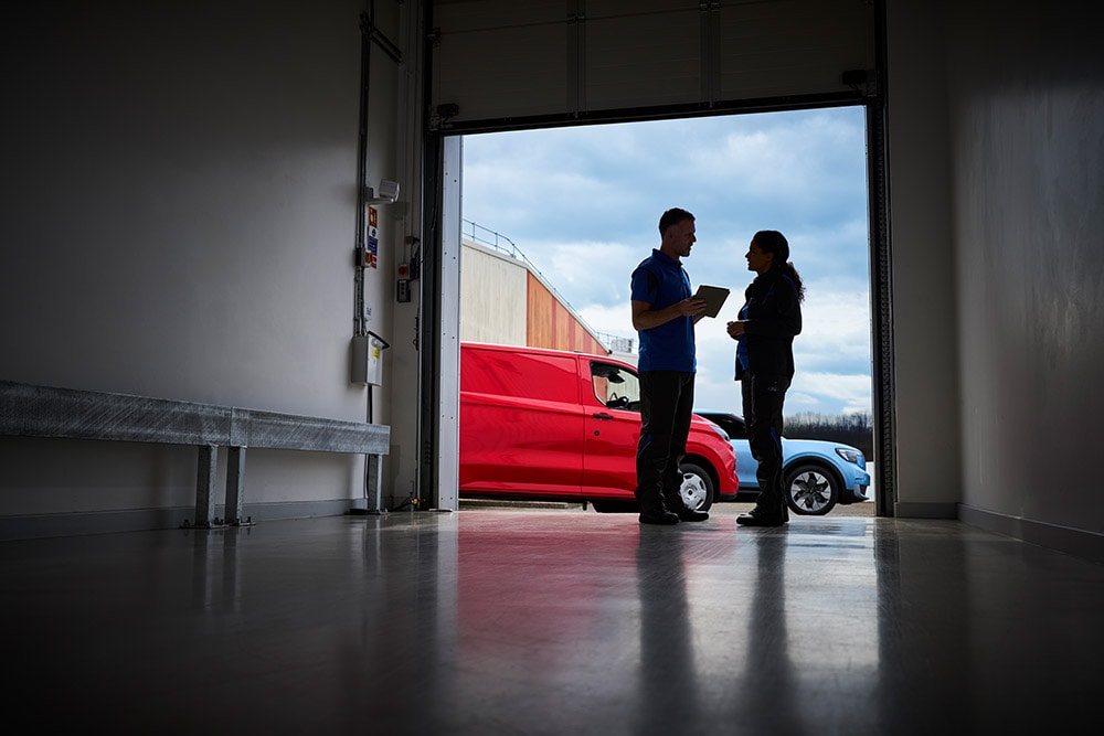 Dos personas hablando en la puerta de un garaje, con una furgoneta Ford roja y un SUV Ford azul estacionados fuera.
