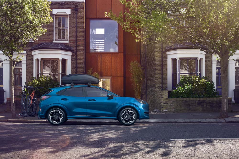 Vista lateral de un Ford Puma azul con una caja para equipaje en el techo y un portabicicletas, estacionado en una calle residencial.