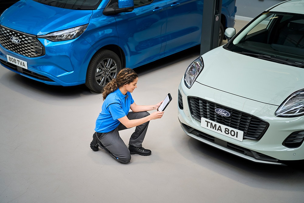 Un técnico mira una tablet arrodillado entre una camioneta Ford azul y un Ford Puma blanco.