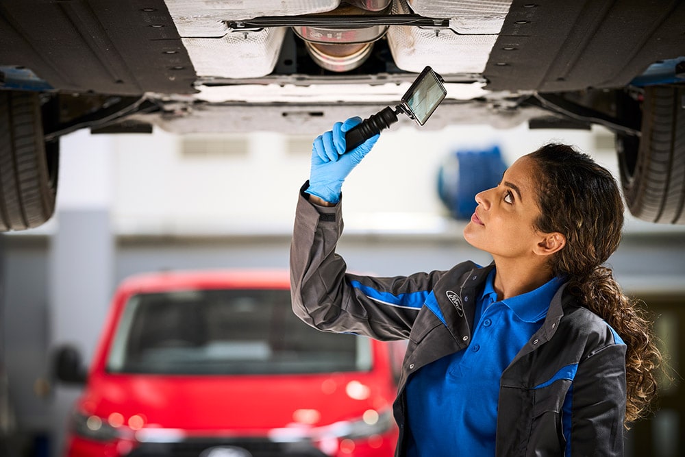 Un mecánico de Ford inspeccionando los bajos de un vehículo, con una furgoneta Ford roja aparcada al fondo.