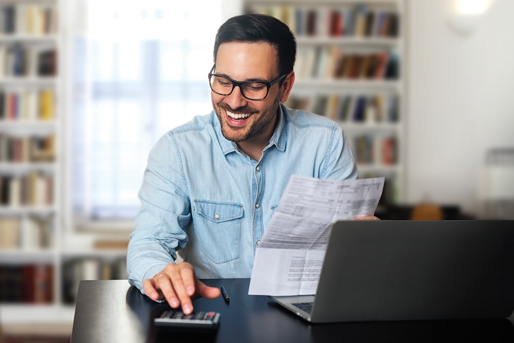  Un hombre con gafas sonríe mientras usa una calculadora y sostiene documentos en un escritorio con un portátil.