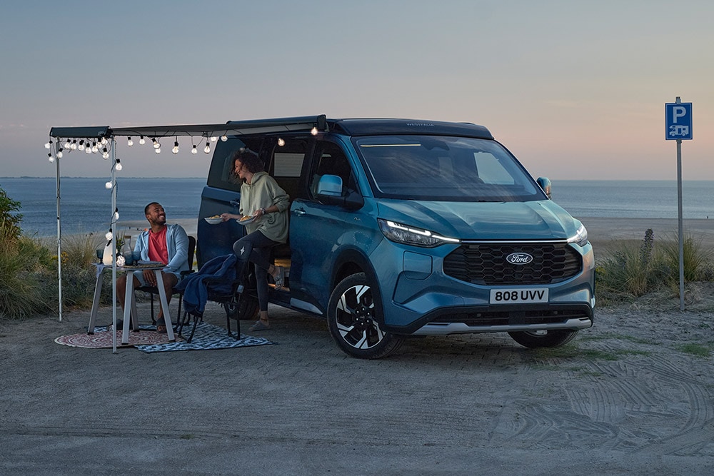 Dos campistas relajándose junto a un Ford Pro Transit Custom Nugget azul, con una zona de aparcamiento junto a la playa al fondo.