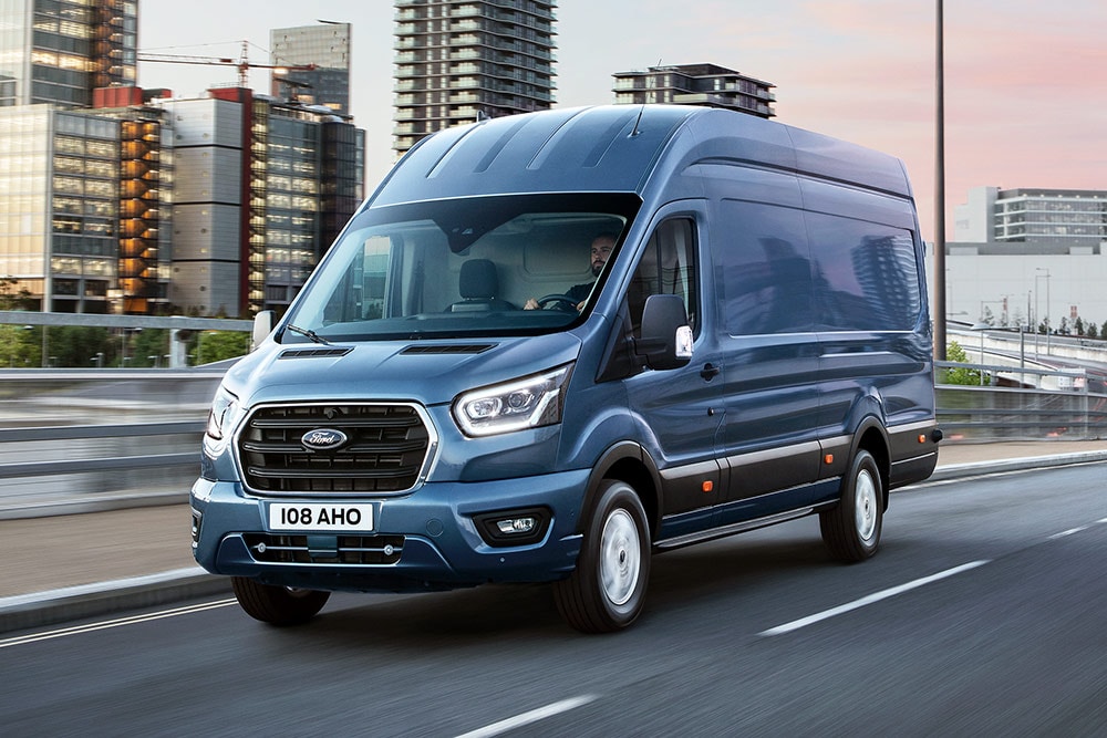 Un Ford Transit plateado circulando por un paisaje urbano.