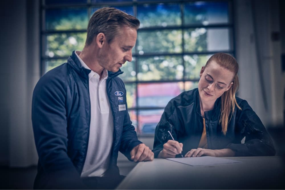 Dos empleados de Ford Pro en una mesa, uno firmando un documento, el otro observando en un taller.