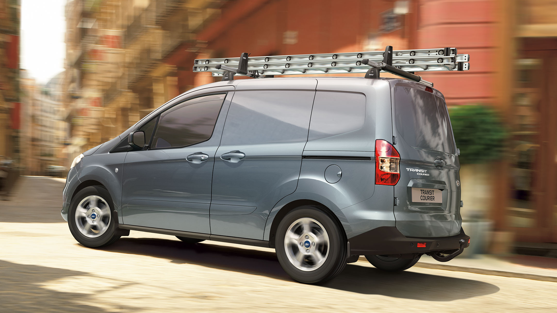 Ford Transit Courier de color azul plata parada en la carretera con portaequipajes y escalera. Vista de tres cuartos trasera