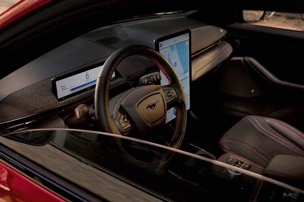 Interior of a Ford Mustang Mach-E, showing the steering wheel, digital instrument cluster, and large central touchscreen. 
